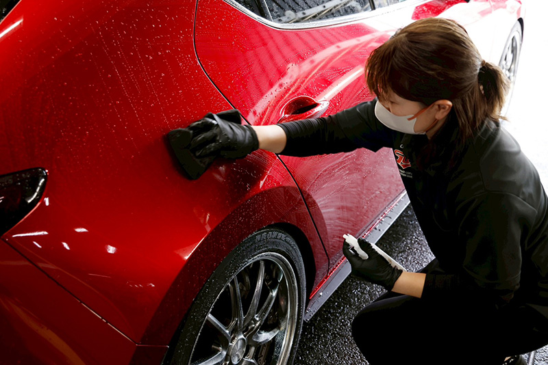 赤い車のボディにコーティングメンテナンスを行う作業員が、洗車後に専用スポンジで丁寧に拭き上げている様子